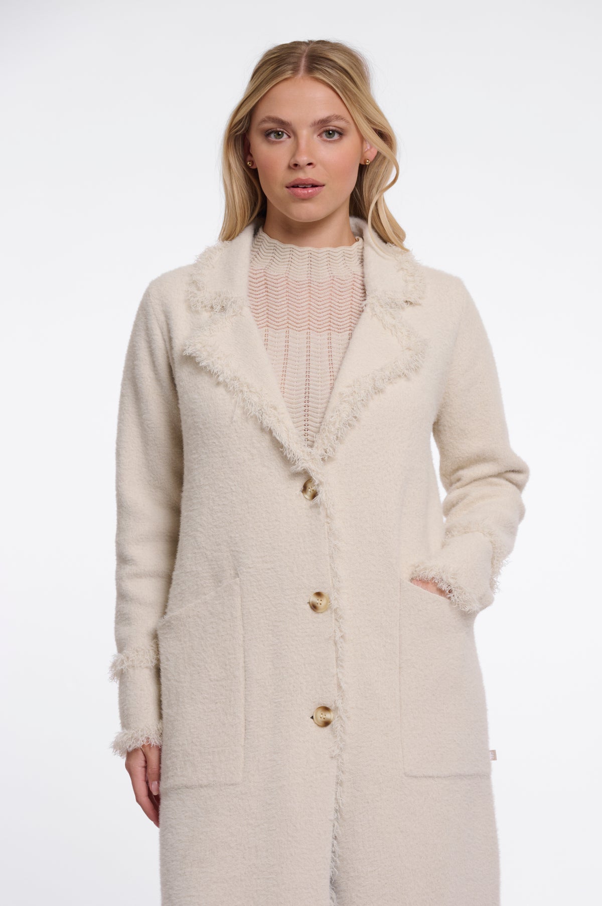 A woman wearing a cream-colored textured coat with frayed trim and gold buttons, layered over a pink ribbed top, against a plain white studio background.