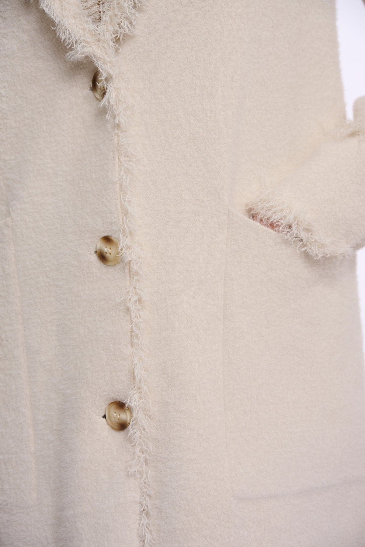 Close-up of a cream-colored textured jacket featuring frayed fringe trim and metallic gold buttons.