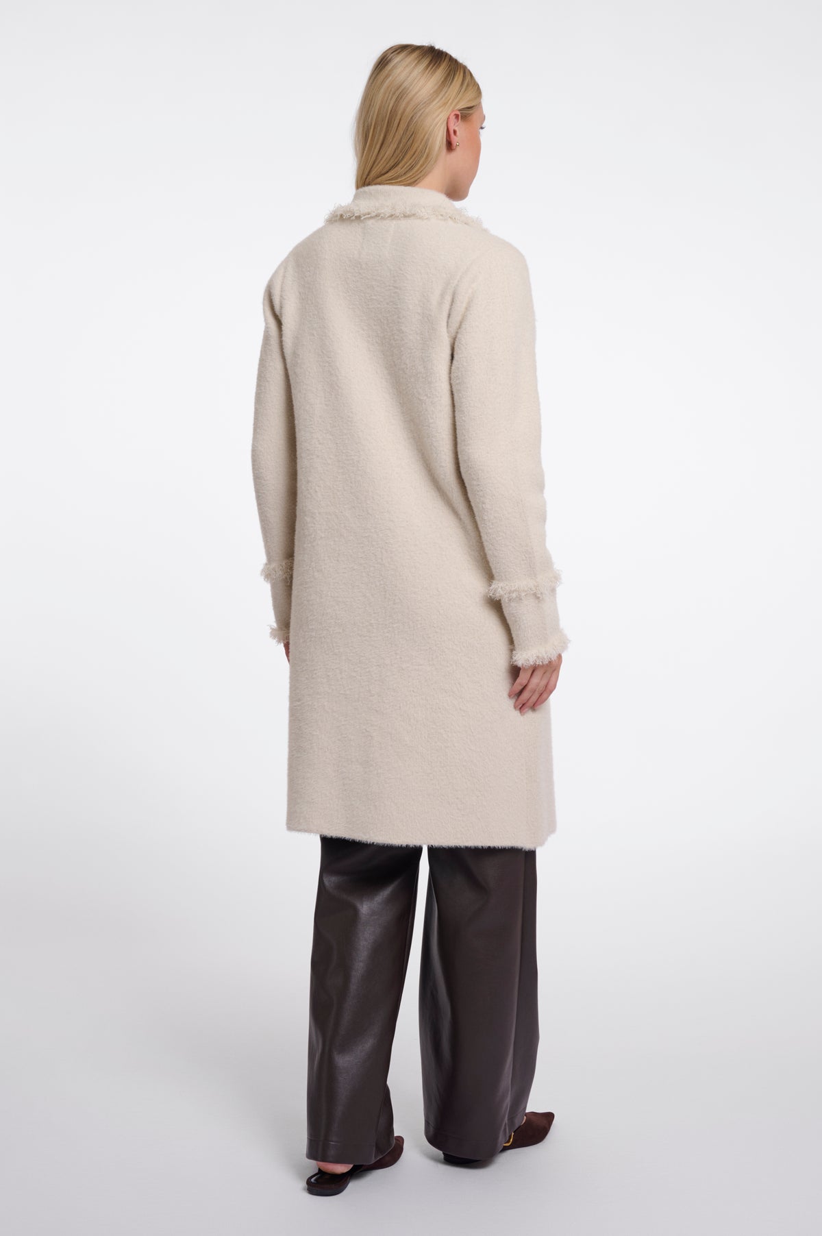 A woman shown from the back wearing a long cream-colored fuzzy coat with frayed trim, paired with dark brown wide-leg pants and black shoes, against a plain white studio background.