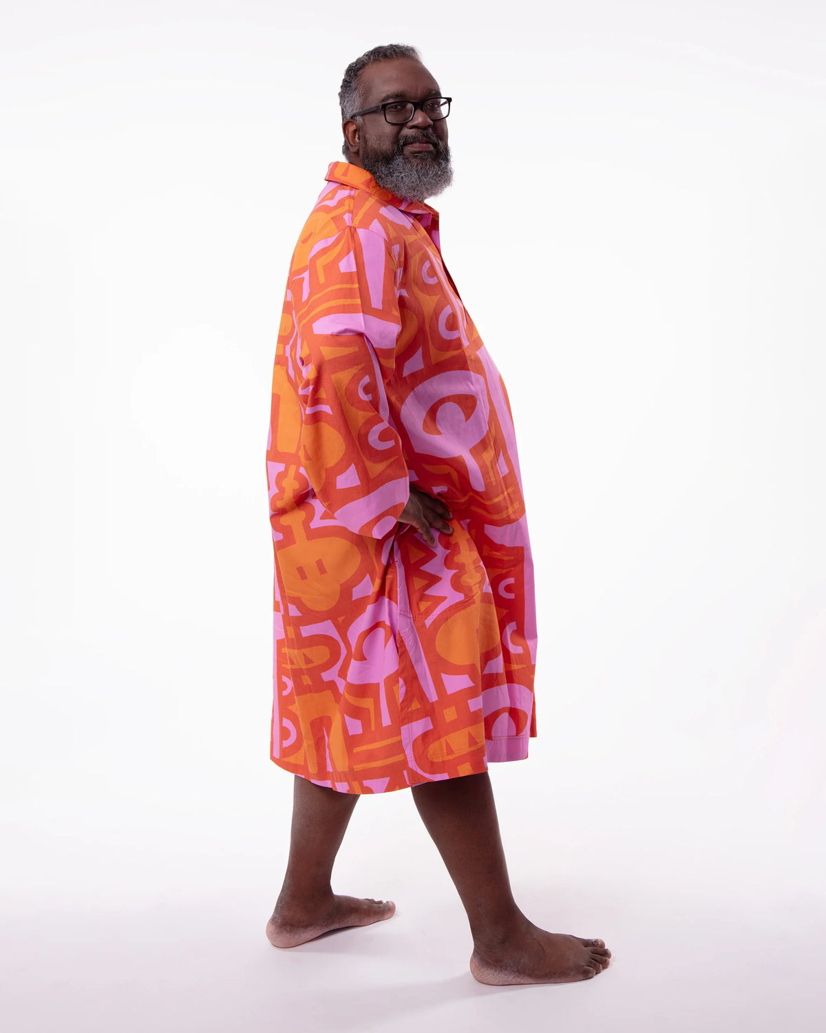 A man wearing a vibrant orange and pink abstract patterned robe, standing barefoot against a plain white studio background.