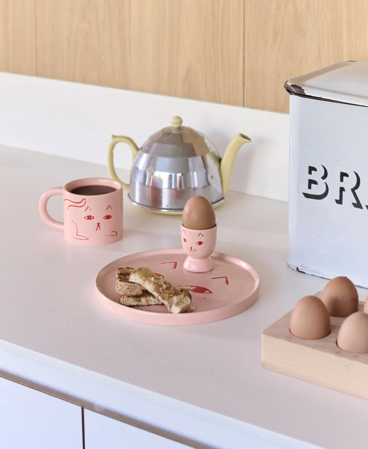 Pink egg holder with a cracked egg on a plate, mug, teapot, and eggs on a kitchen counter.
