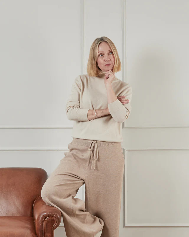 Woman in a beige sweater and light beige pants from Chalk UK, posing thoughtfully next to a brown leather sofa.