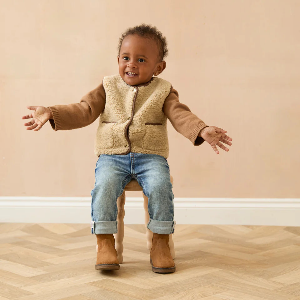 Child wearing a beige vest, brown sweater, blue jeans, and brown boots on a wooden floor.