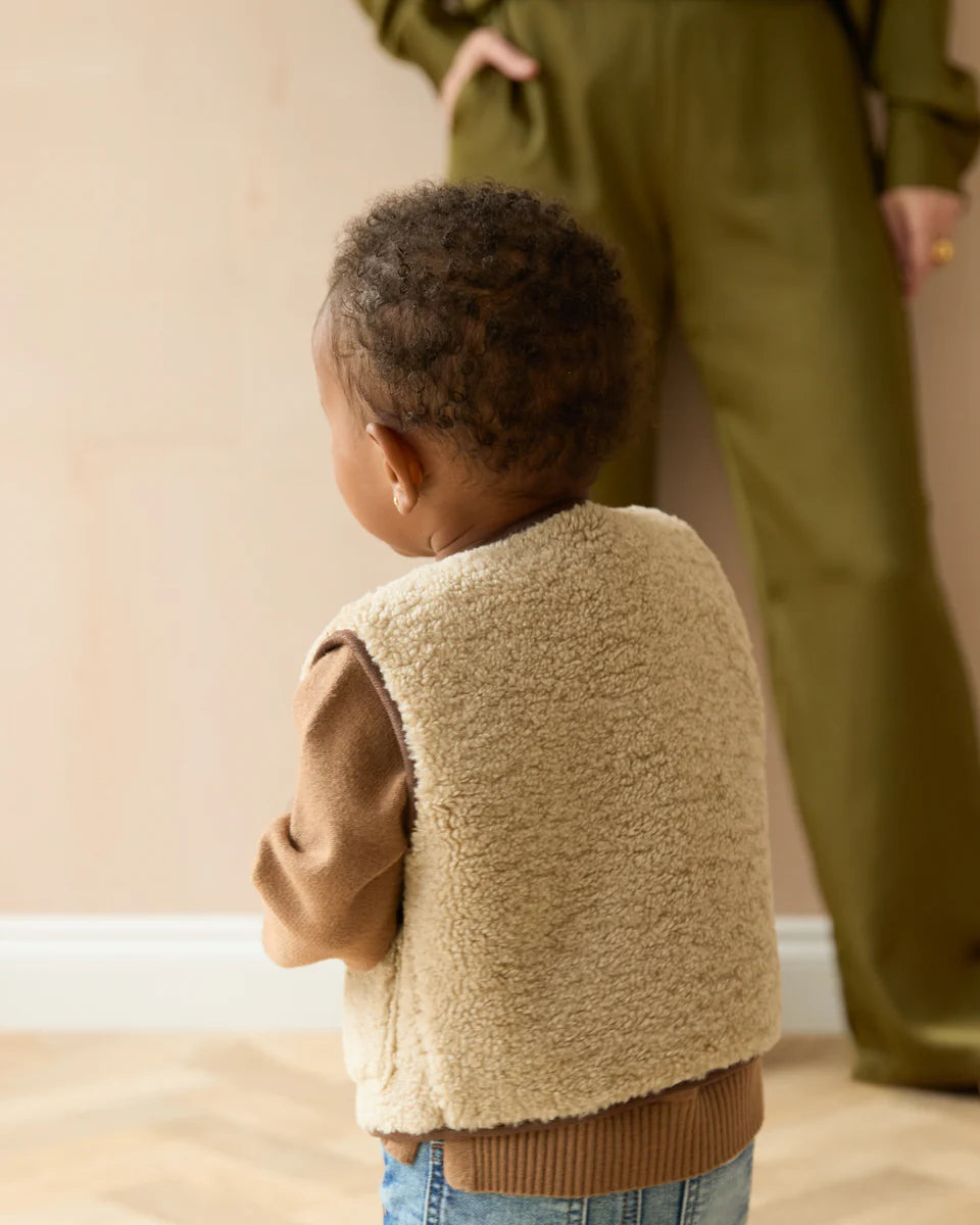 Child wearing a beige fleece vest standing next to an adult in a green outfit.