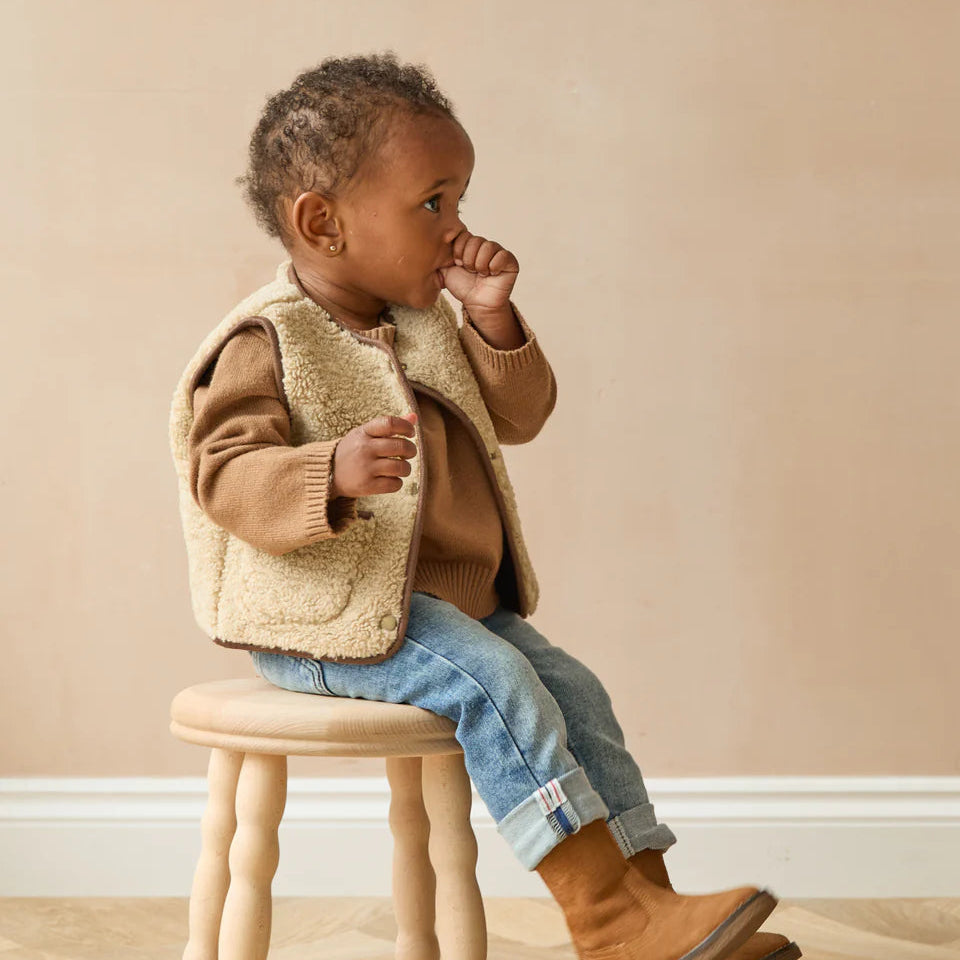 Child sitting on a small stool wearing a brown sweater, beige vest, blue jeans, and brown boots against a beige wall.