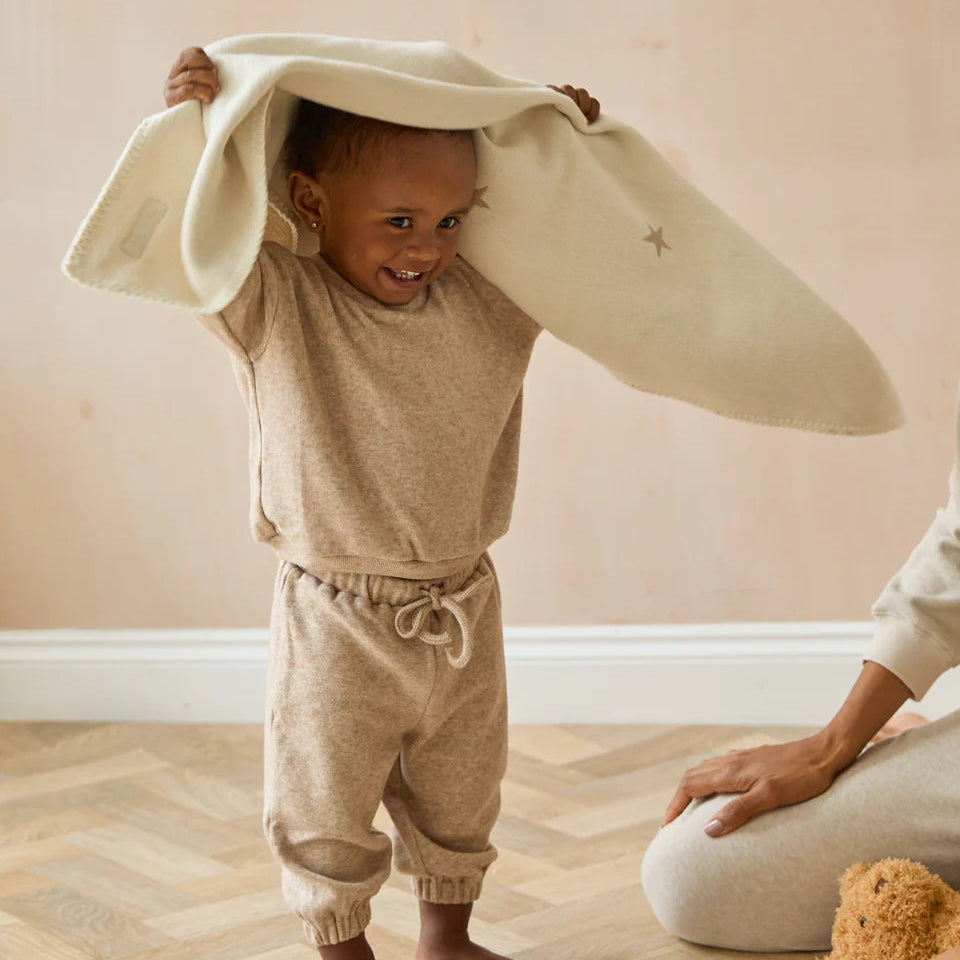 Child wearing a beige outfit with a large white hat, standing on a wooden floor.