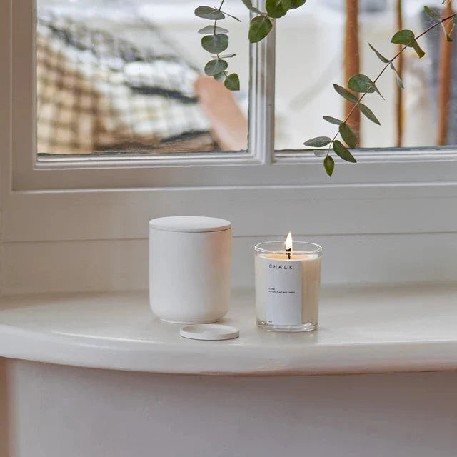 A lit Chalk candle in a clear glass jar and a white ceramic candle container with lid.