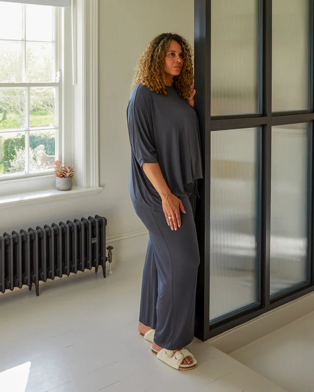 A woman stands in a room wearing Chalk UK’s dark grey lounge set and cozy light slippers.