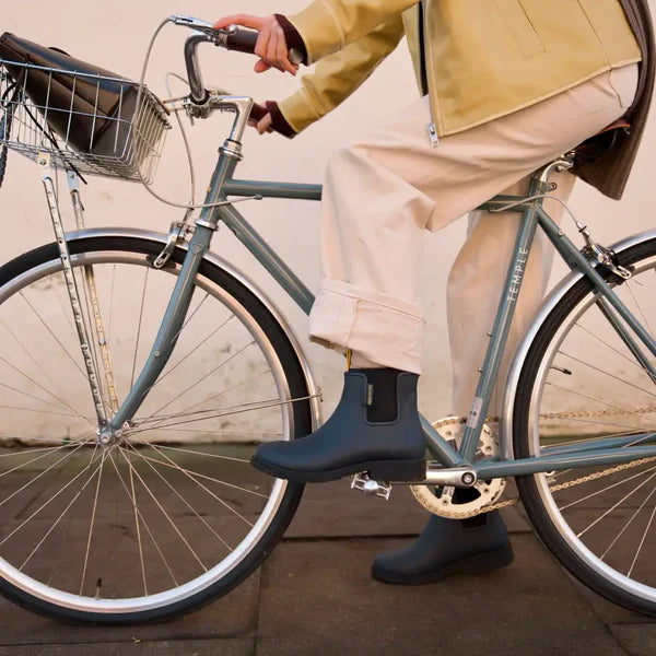 Person wearing navy rain boots standing next to a bicycle on a sidewalk.