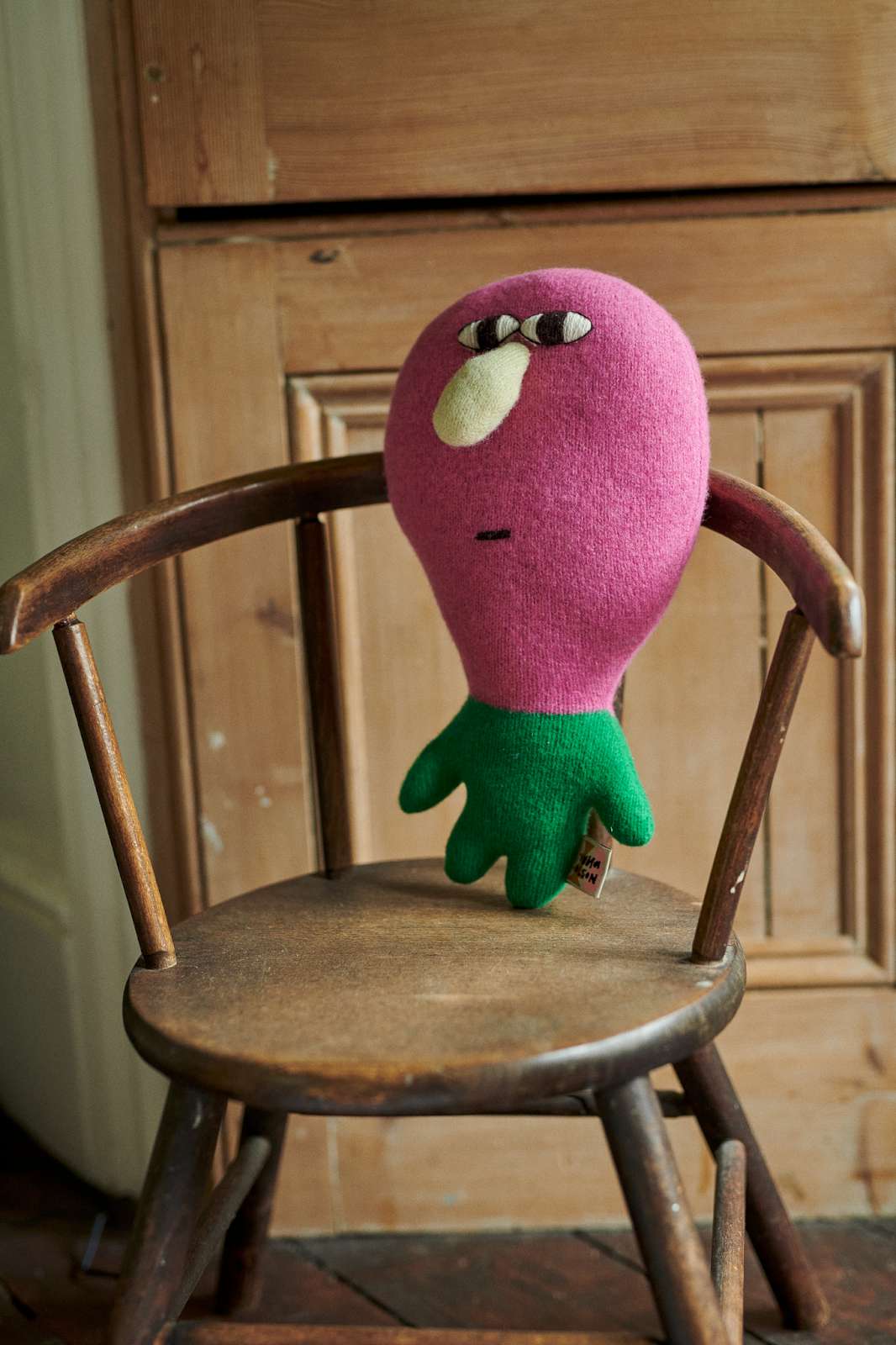 A pink and green plush toy with a face sits on a rustic wooden chair in front of a wooden cabinet.