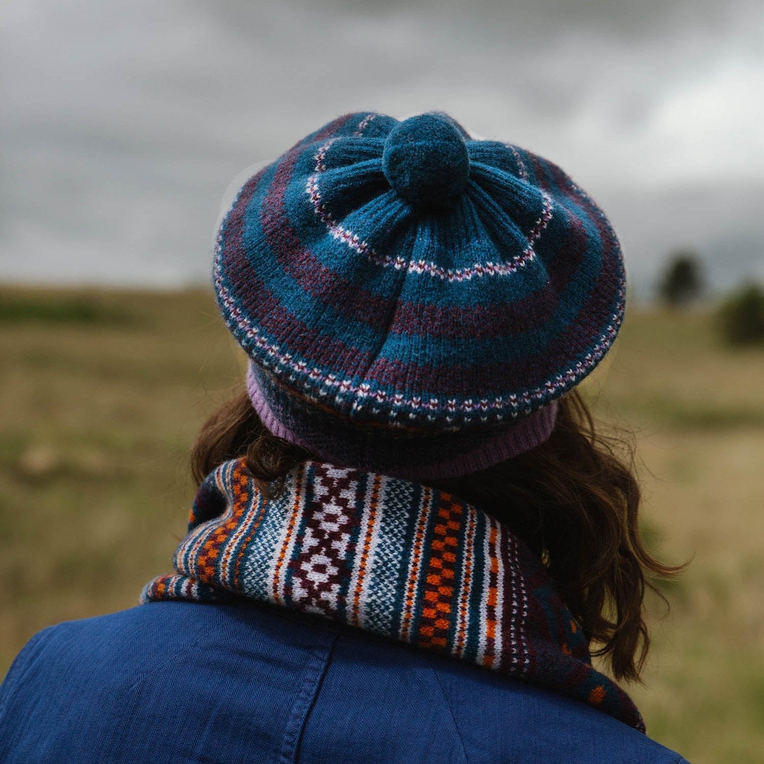 Striped teal and purple knitted beret with a textured top.  The model also wears a multicolored striped scarf.