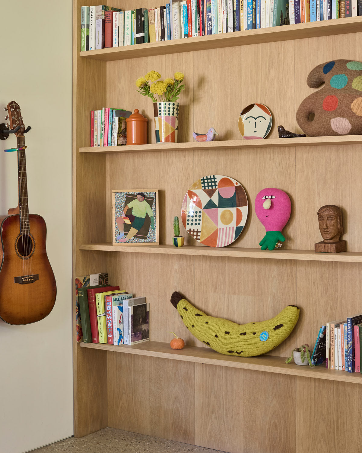 Wooden shelf with books, decorative items, and a guitar against a beige wall.