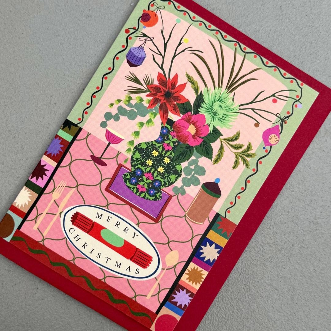 A square Christmas greeting card featuring a colorful floral arrangement, a pink patterned background, and a 'Merry Christmas' oval label, photographed on a neutral gray surface.