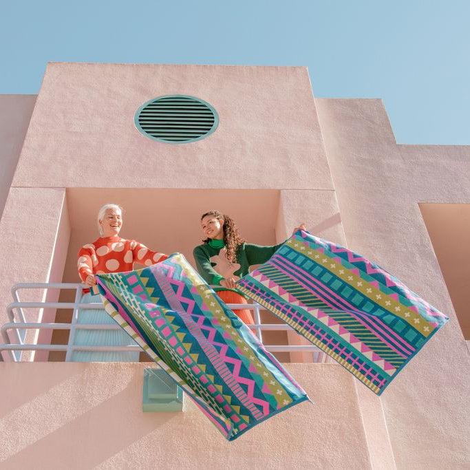 Two people standing on a pink stucco balcony under a clear blue sky, holding up two large rectangular beach towels with bright geometric patterns in teal, pink, green, yellow, and blue.