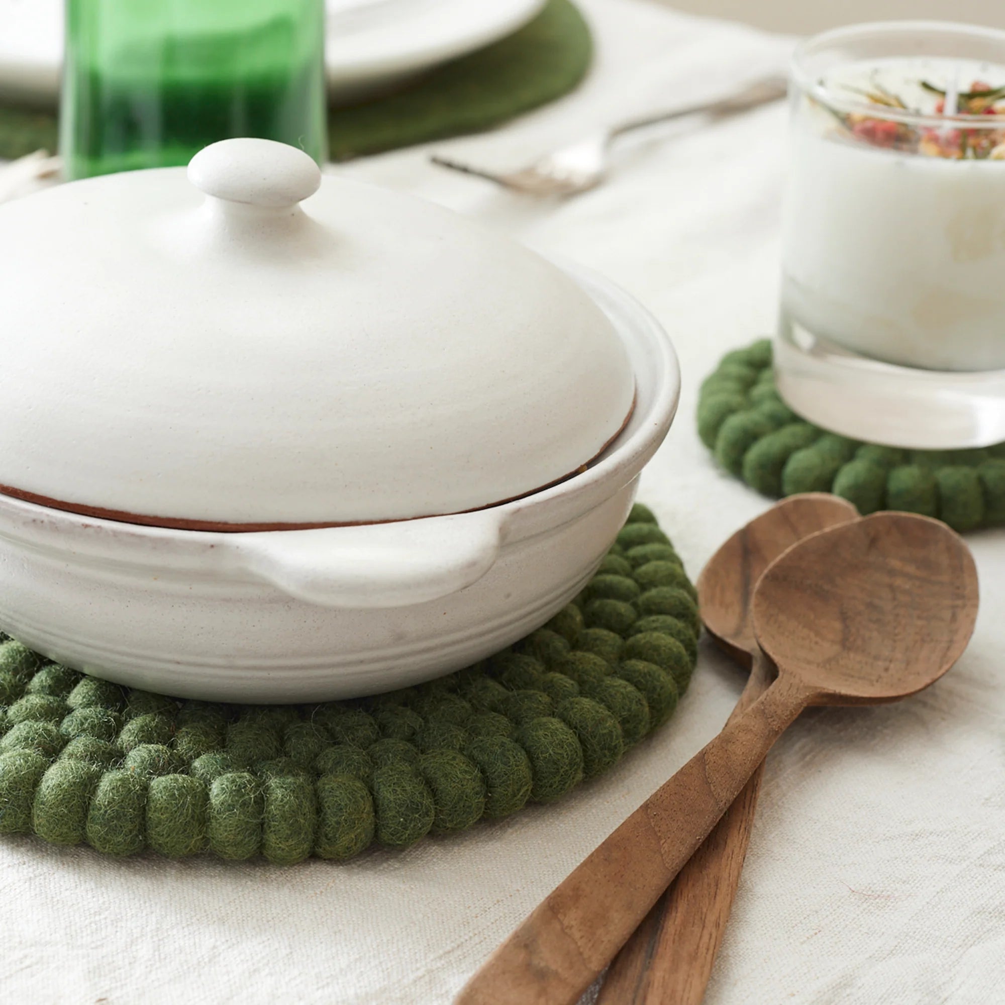 White ceramic pot with lid, resting on a green felt coaster.  Two wooden spoons are also visible.