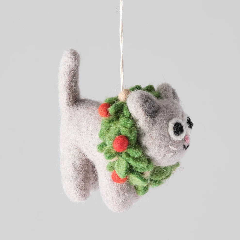 A gray felted wool cat ornament hanging from a white string, decorated with a green wreath and red berries, set against a solid light gray background.