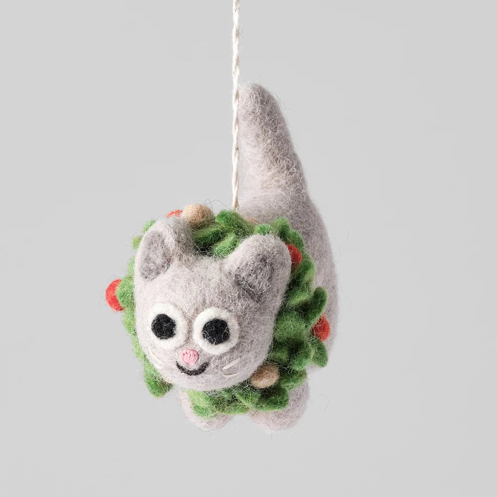 A felted wool cat ornament hanging from a white string, with a green wreath-like body, red and beige accents, and large black-and-white eyes, shown against a plain light gray background.