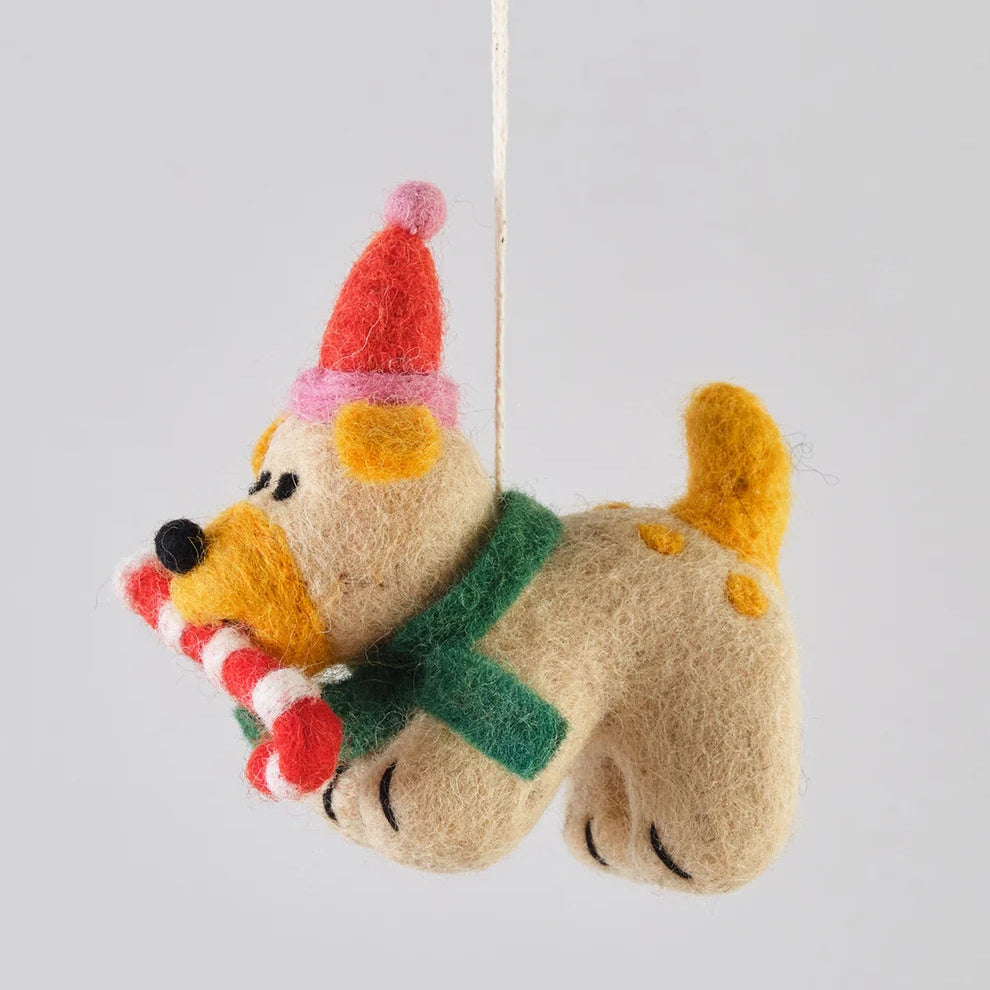 A felt dog Christmas ornament hanging from a string, wearing a red Santa hat, green scarf, and holding a striped candy cane, set against a plain light gray background.