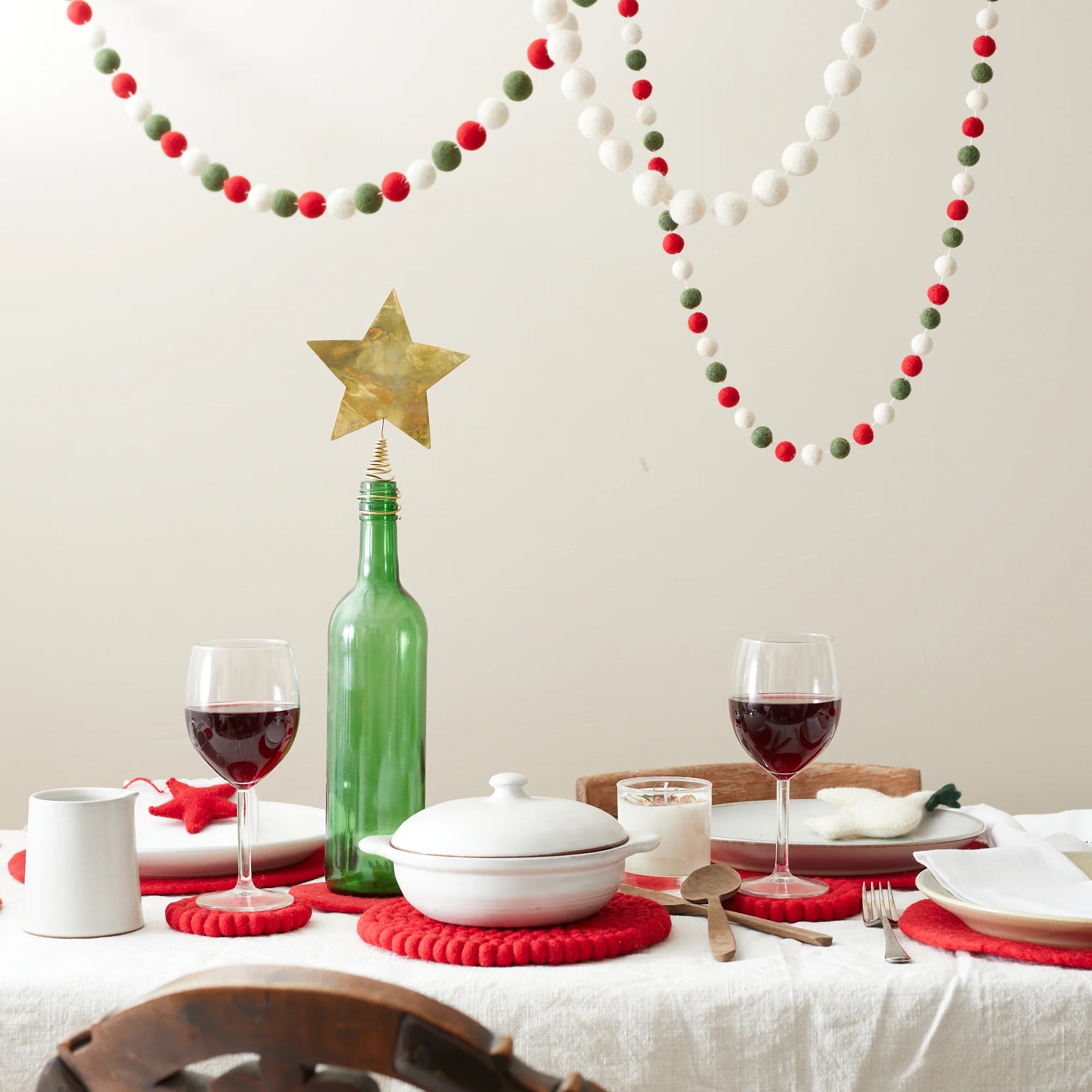 Christmas table setting with red felt coasters, a white casserole dish, wine glasses, and a wine bottle topped with a gold
