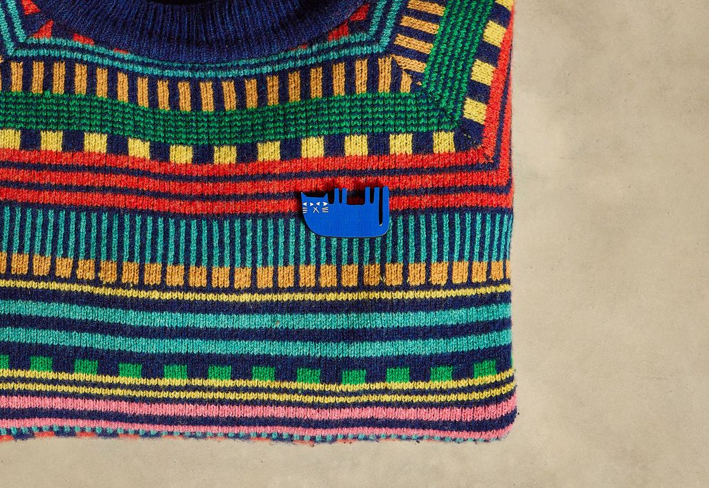 A close-up of a colorful hand-knit sweater featuring a geometric pattern in green, blue, red, yellow, and pink, with a small blue whale-shaped pin on the chest, set against a neutral beige background.