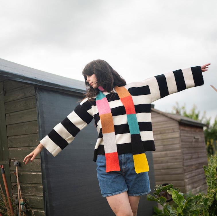 A woman in a black and white striped sweater and denim shorts poses, wearing a multi-colored scarf by Donna Wilson.