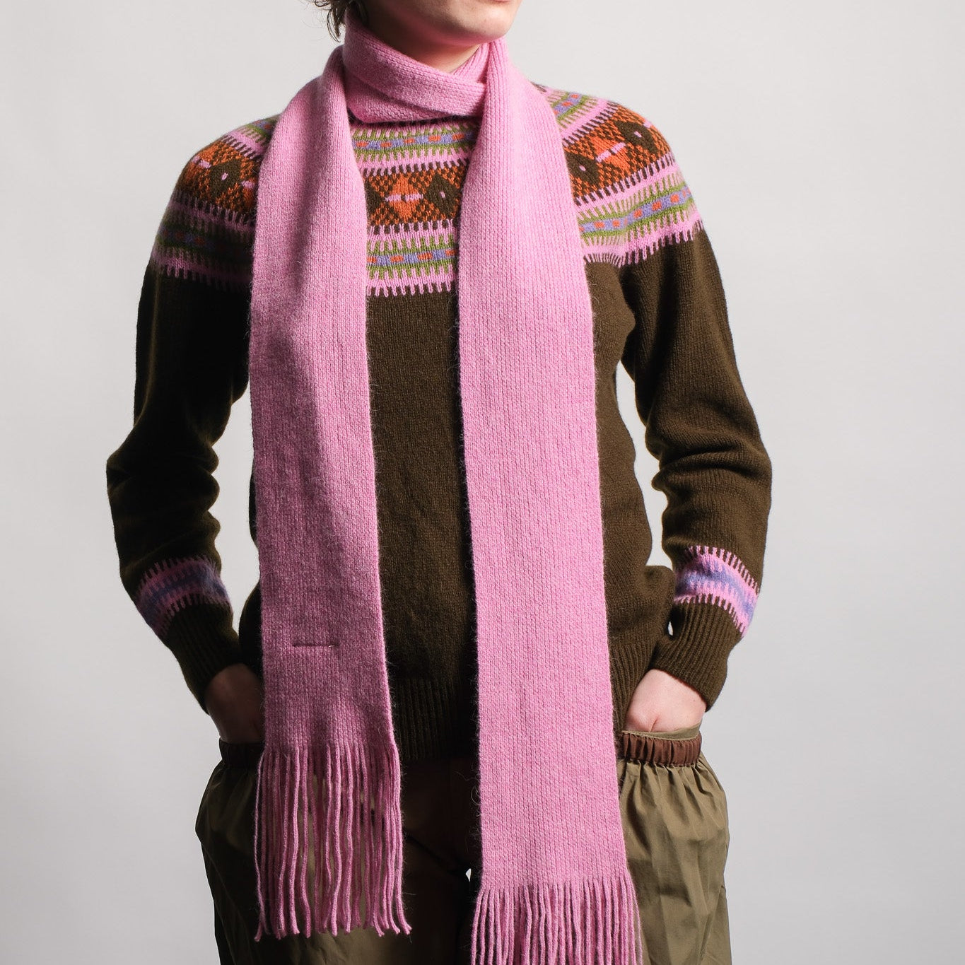 A model wearing a long, soft pink knit scarf with fringe ends, styled over a brown Fair Isle sweater and olive green pants against a light gray studio background.