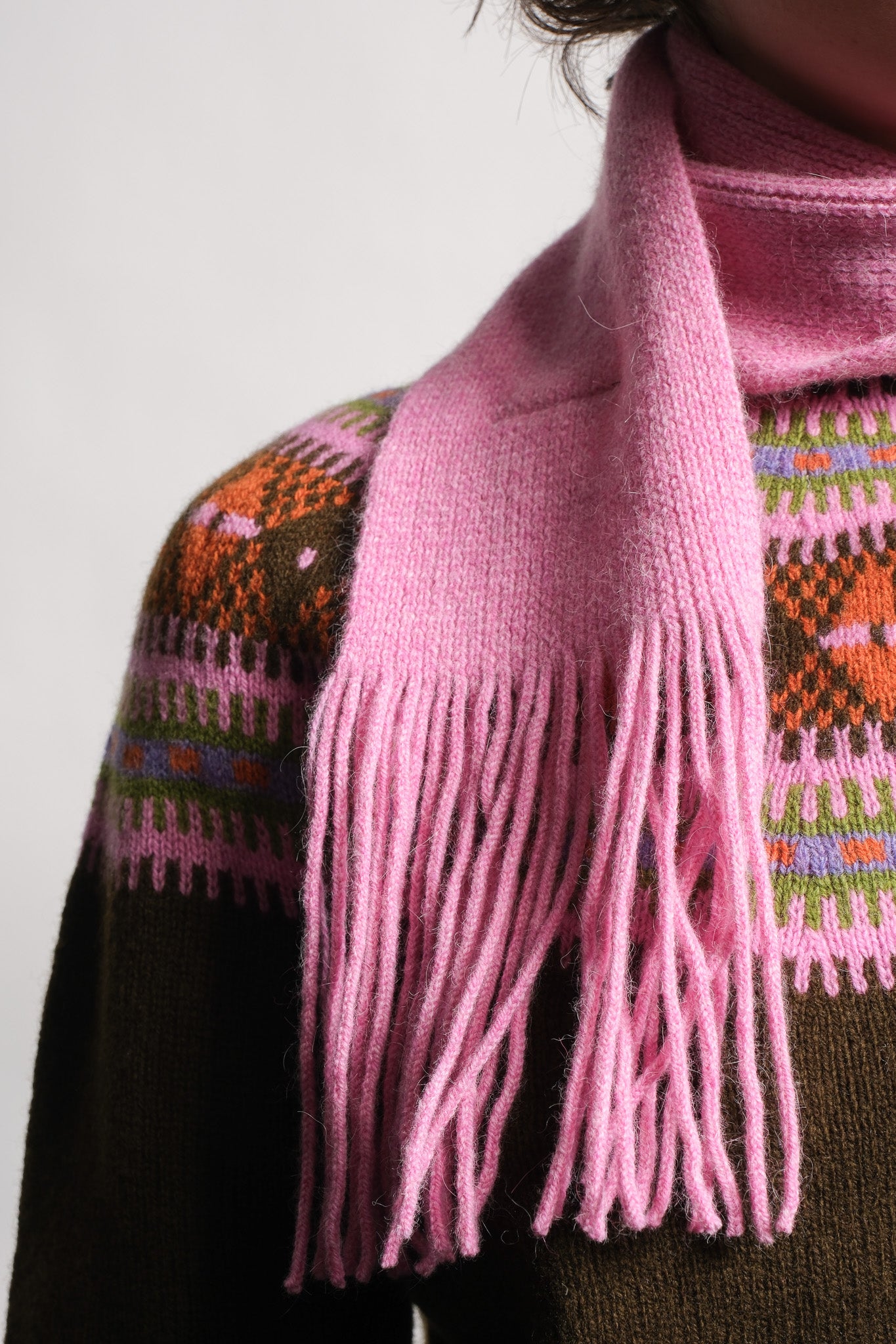 A close-up of a person wearing a soft pink knit scarf with long fringe, layered over a dark brown knit sweater with a colorful geometric pattern.