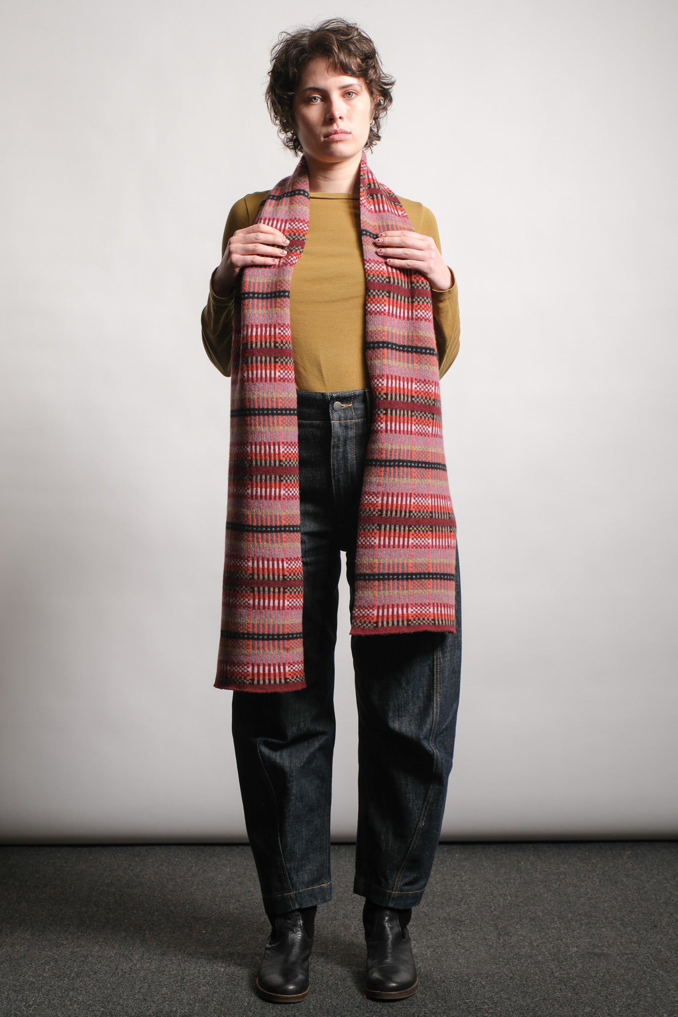 A model stands against a light gray studio background wearing a long, wide striped scarf in red, pink, orange, and dark brown tones over a mustard yellow top, dark blue jeans, and black boots.