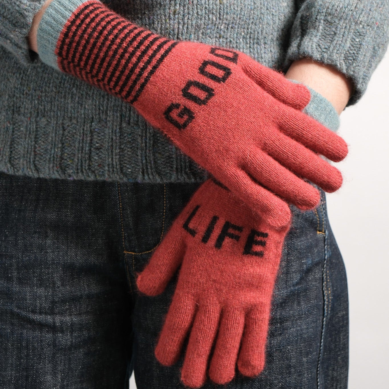 A person wearing a gray knit sweater and dark blue jeans, holding a pair of coral-red knit gloves with black text reading 'GOOD LIFE' and black striped cuffs.
