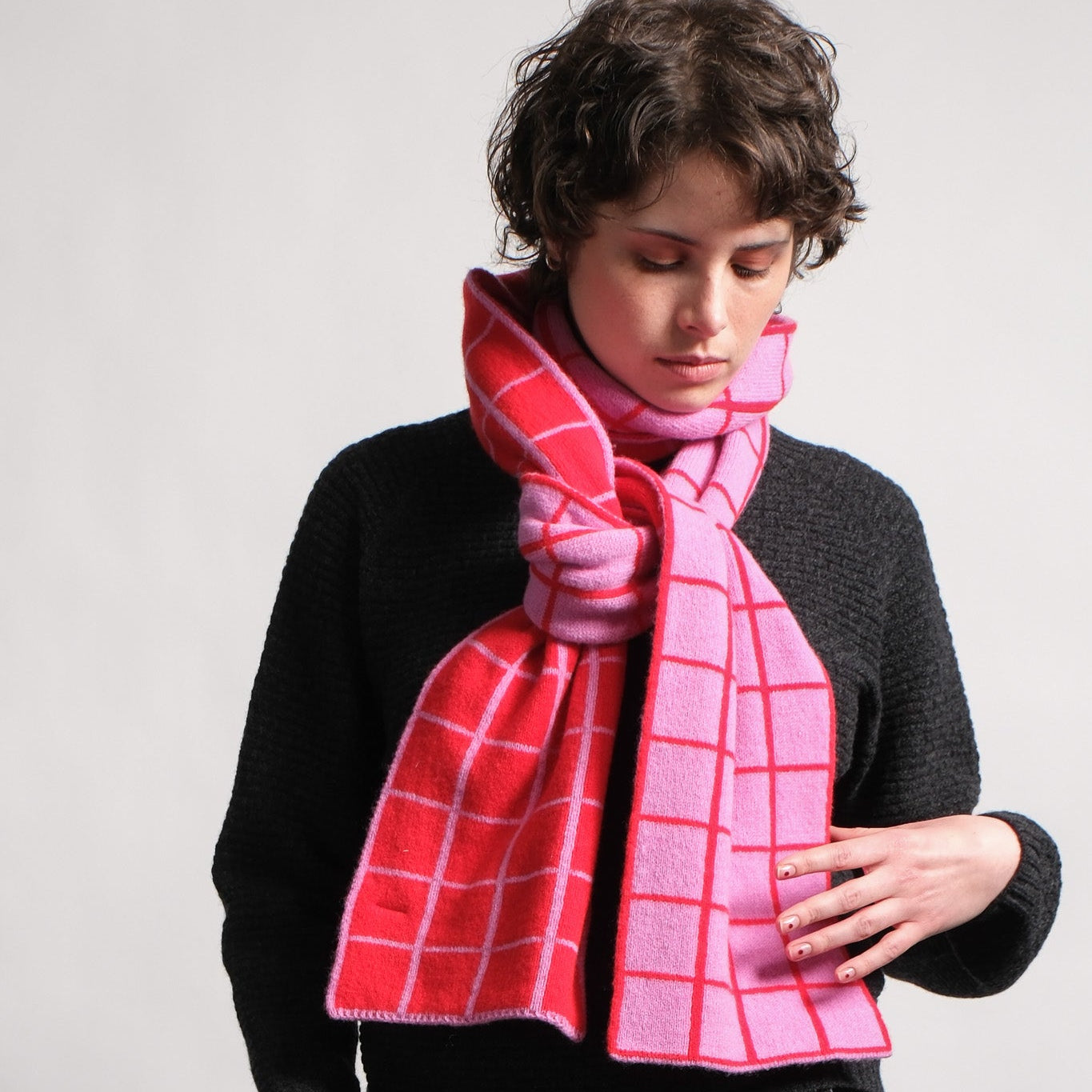 A model wearing a pink and red plaid scarf with white grid lines, styled over a black sweater and dark jeans against a light gray background.