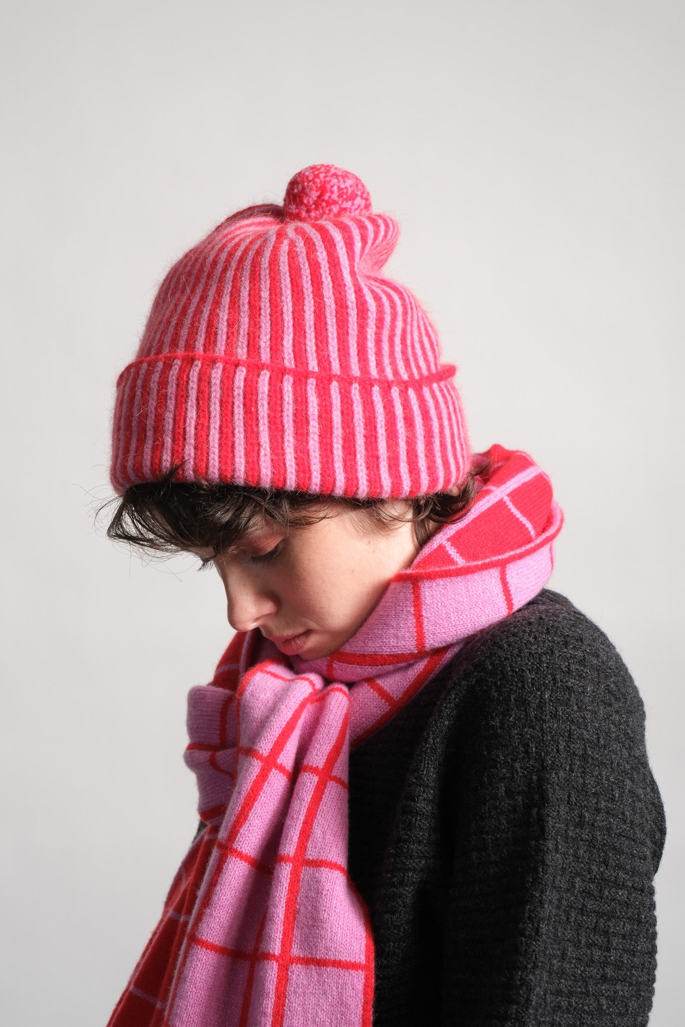 A child wearing a pink and red striped knit beanie with a pom-pom and a matching pink plaid scarf, shown in profile against a plain light gray background.