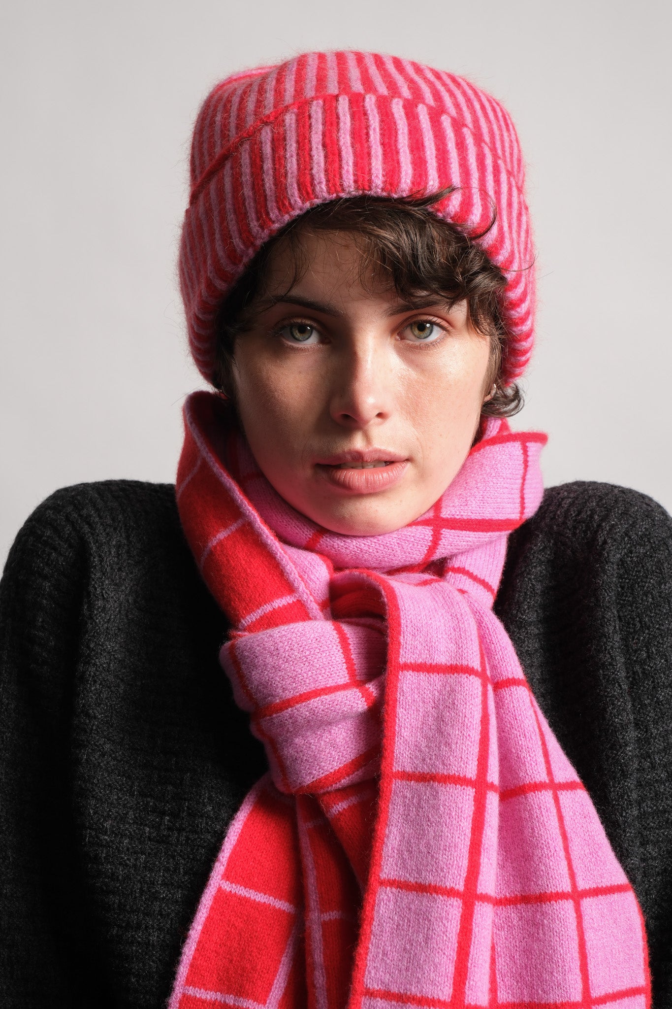 A close-up portrait of a model wearing a pink and red striped knit beanie and a matching pink scarf with a red windowpane pattern, paired with a dark gray knit sweater against a plain light gray background.