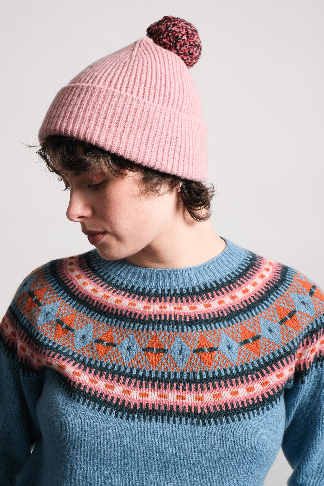 A person wearing a pink ribbed knit beanie with a dark patterned pom-pom and a blue patterned knit sweater, photographed against a plain light gray background.