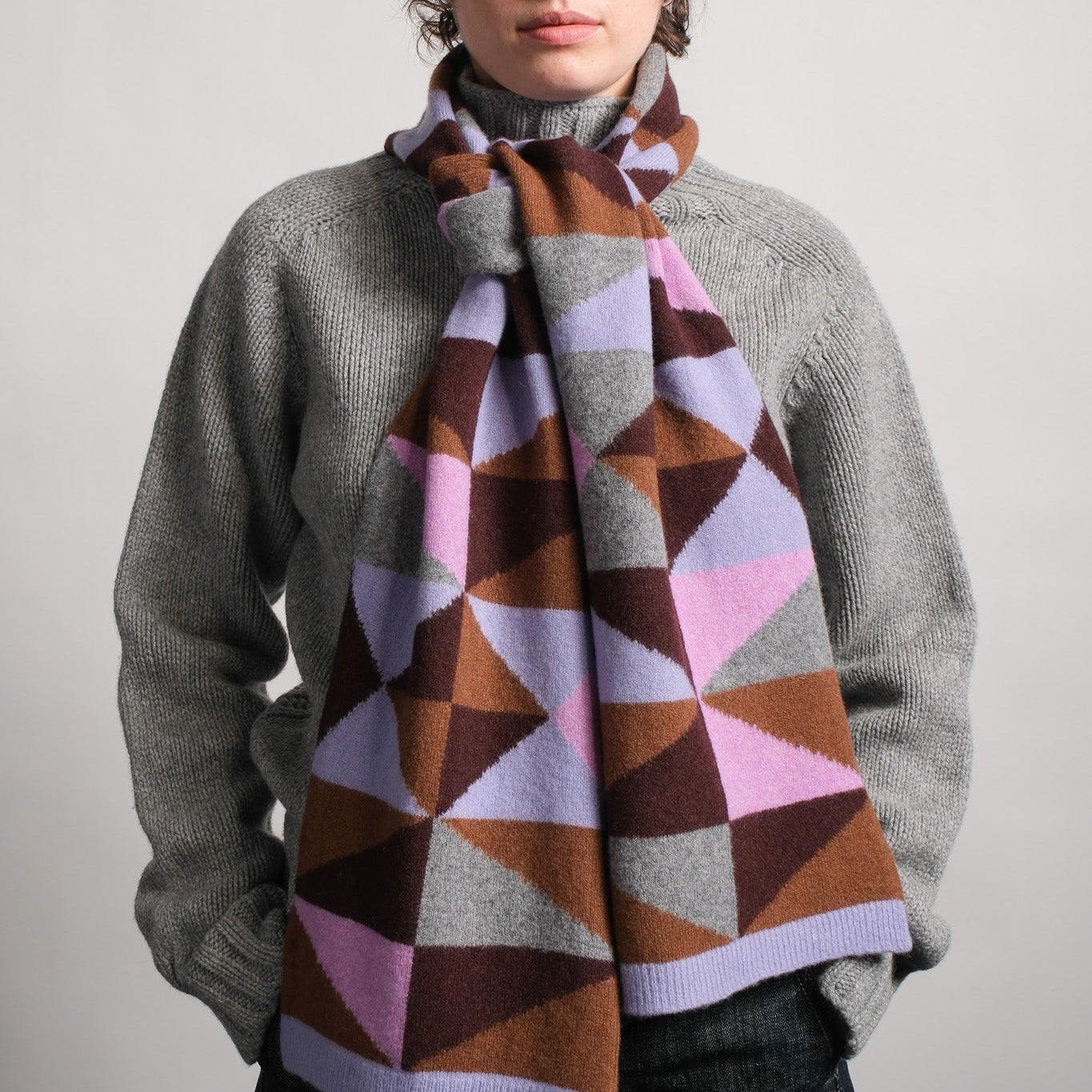 A person wearing a gray knit turtleneck sweater and a thick geometric patterned scarf in brown, pink, purple, and gray, photographed against a plain light gray studio background.