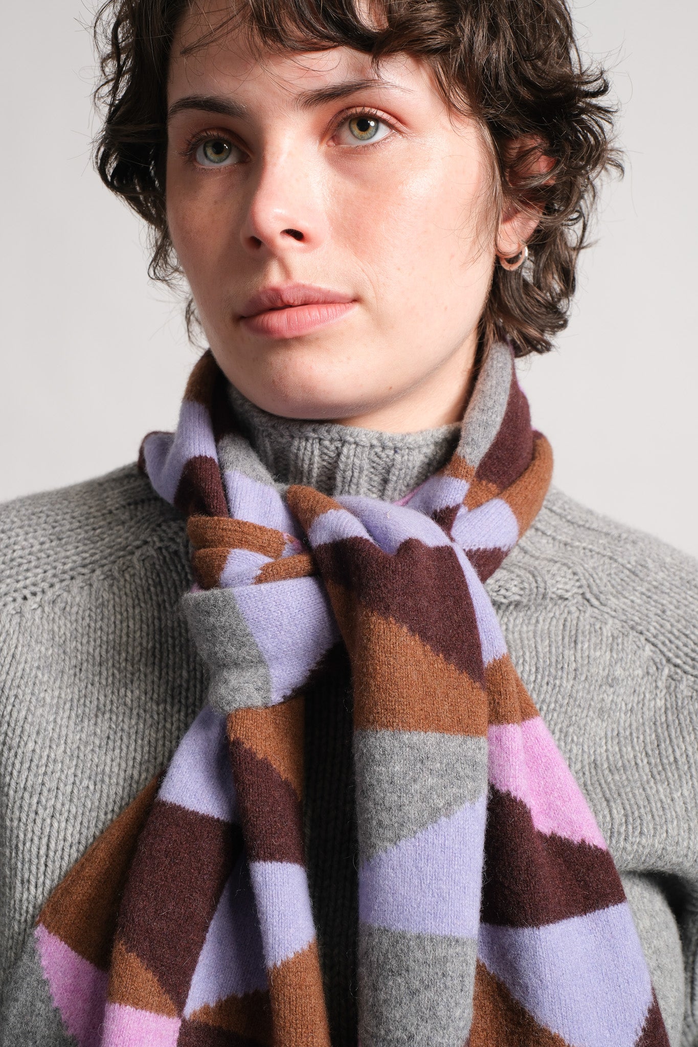 A close-up of a person wearing a chunky knit scarf with a geometric pattern in brown, lavender, gray, and pink over a gray turtleneck sweater.