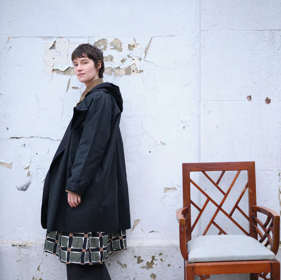 Black hooded coat, knee-length, relaxed fit, worn over a patterned skirt.