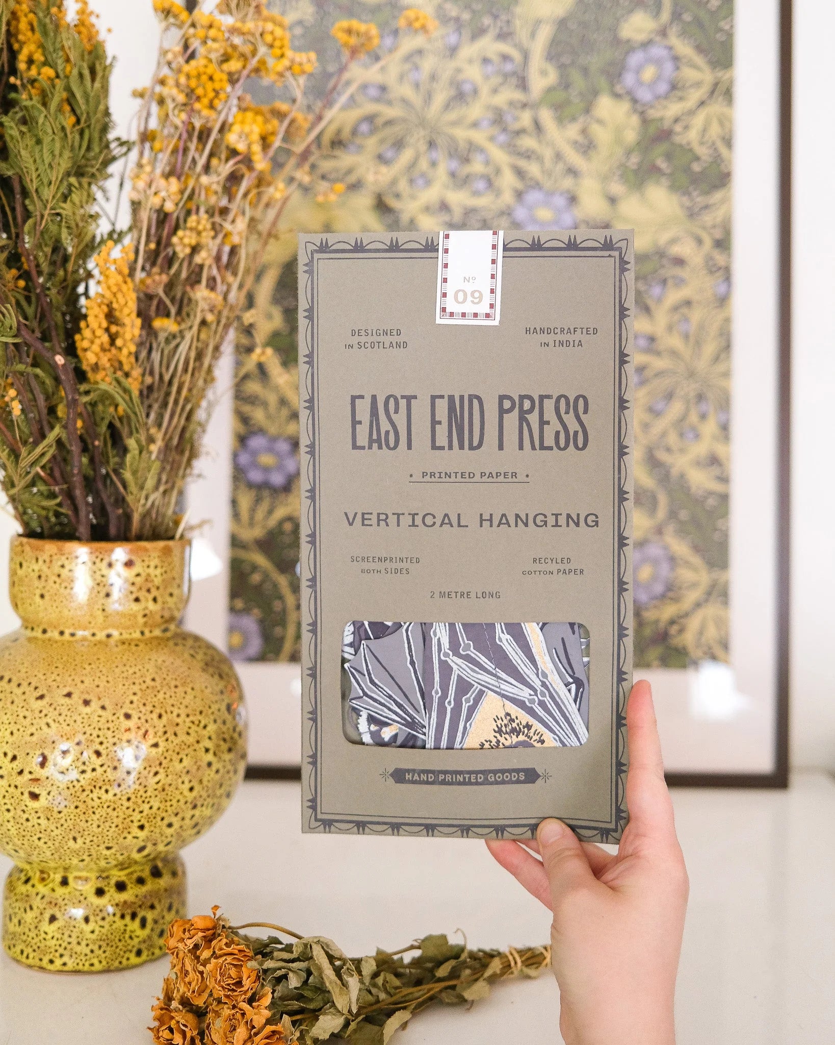 A hand holds a package of East End Press Vertical Hanging printed paper against a warm, styled background featuring a yellow vase with dried flowers and a patterned framed print.