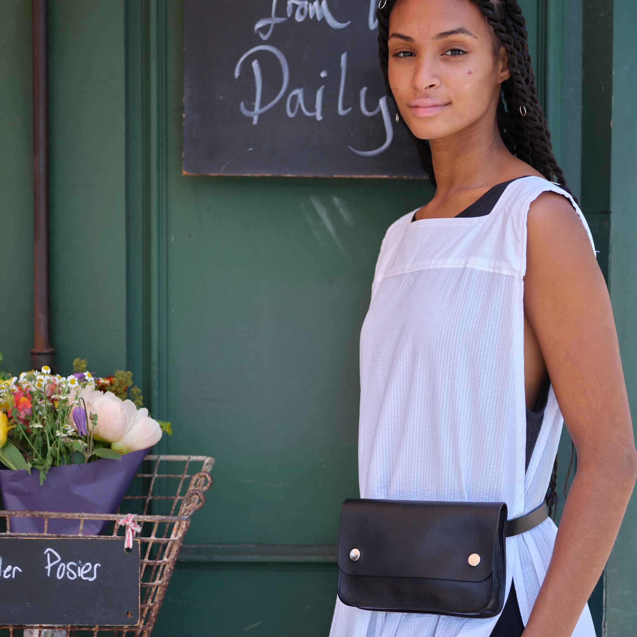 Black leather fanny pack with a silver button closure, worn by a person in a white sleeveless top.