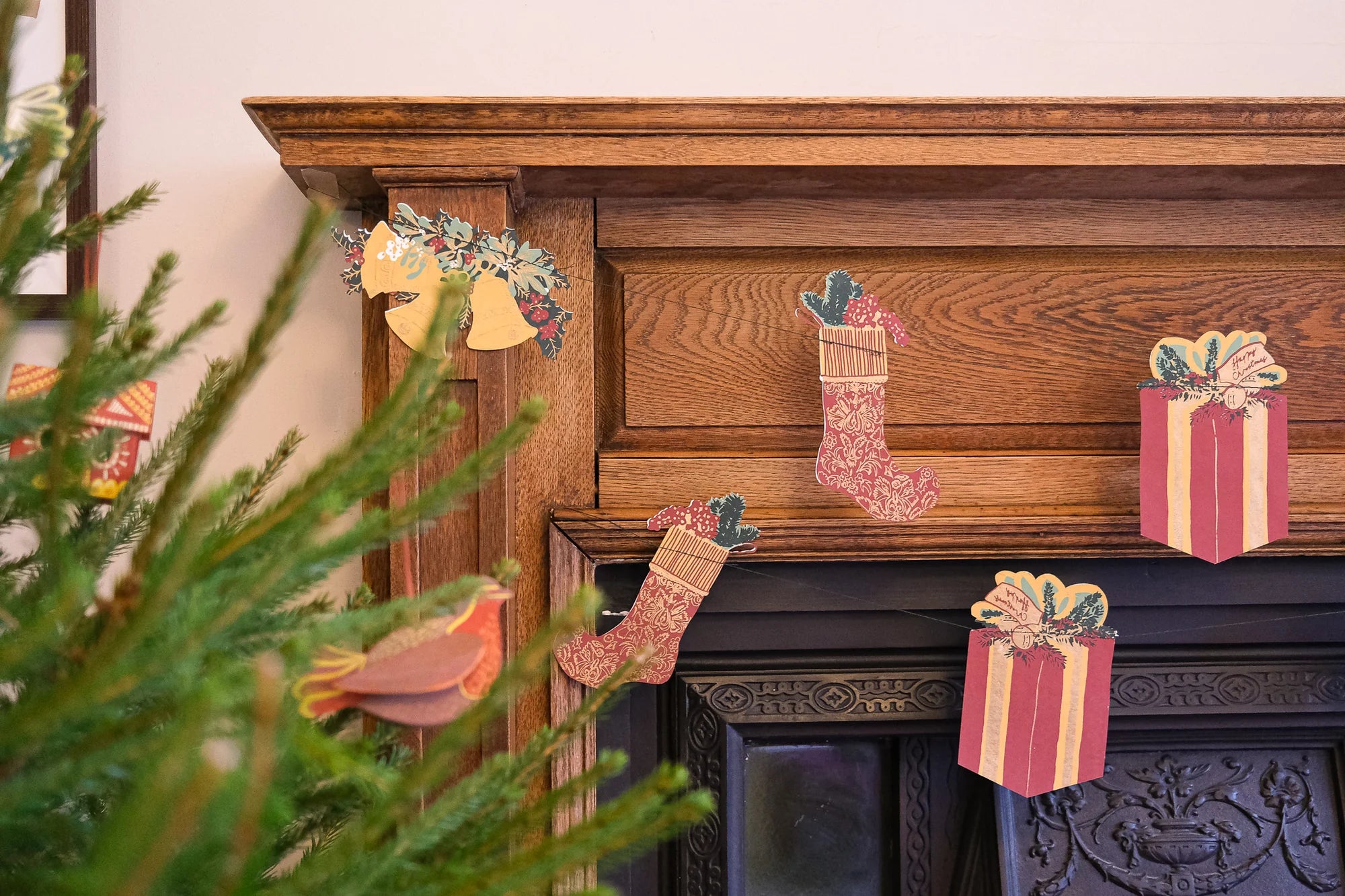 Wooden Christmas mantel decorations including pink patterned stockings, gift boxes, a bird ornament, and a floral wreath, displayed on a dark fireplace with a blurred green pine branch in the foreground.