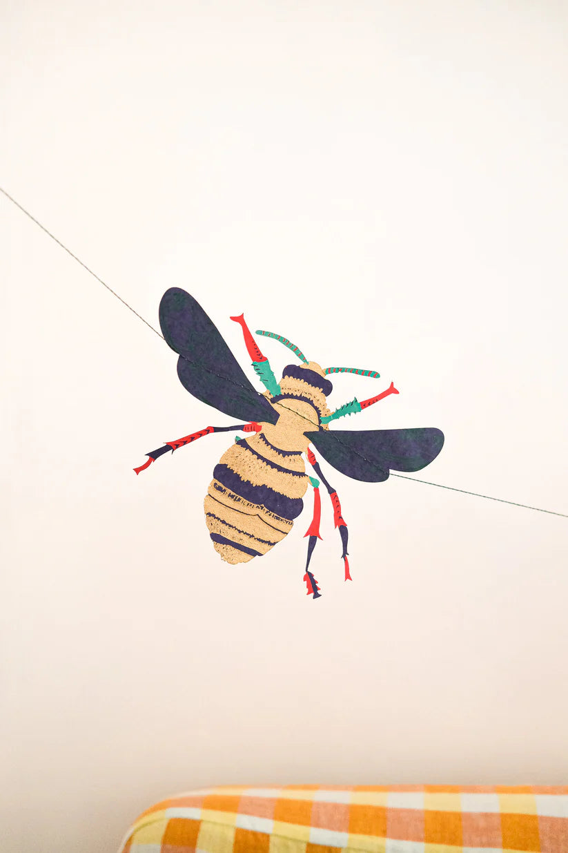 Stylized bee decoration hanging from thin string, gold and dark purple body with red and teal accents.