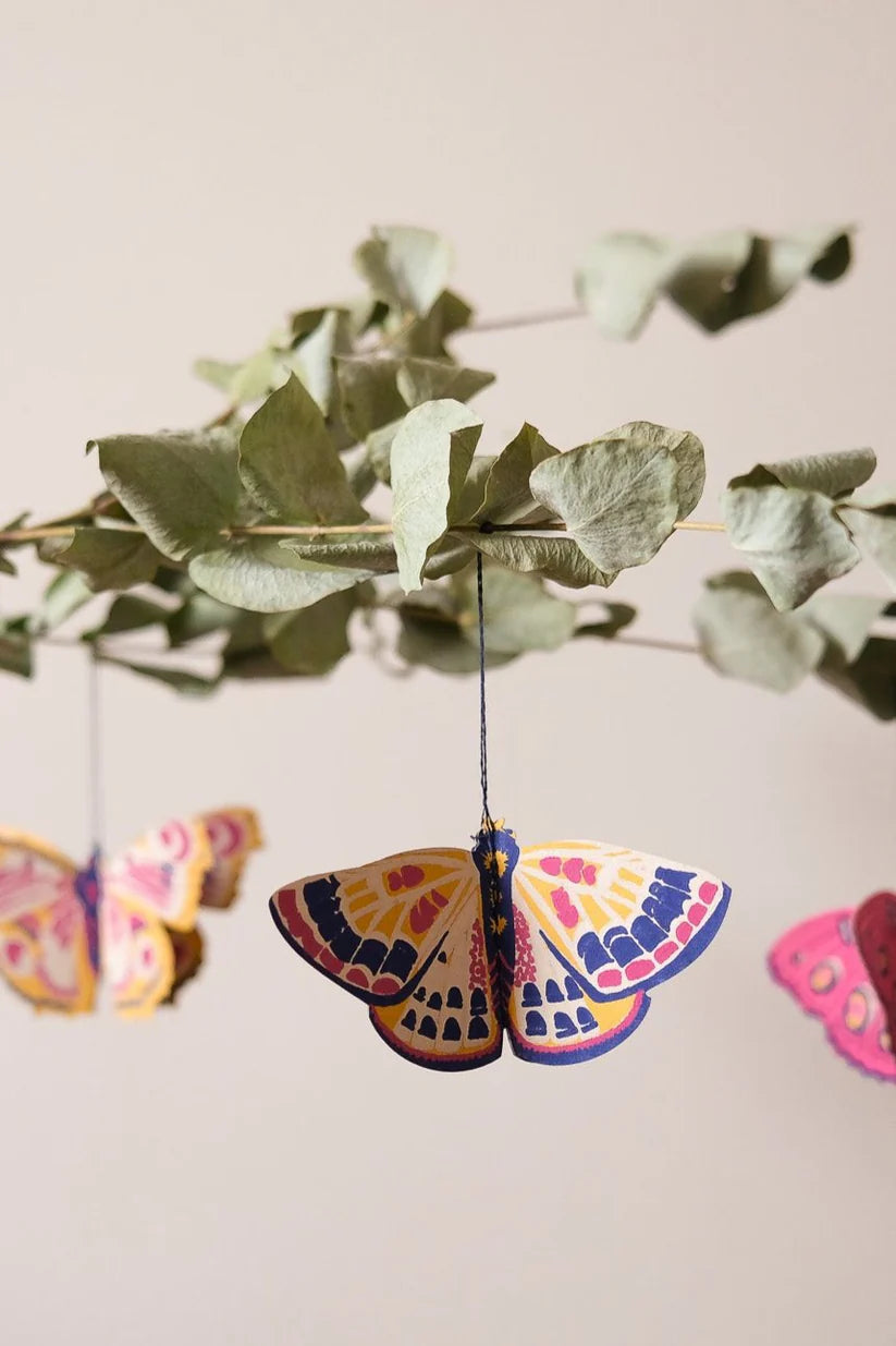 Colorful paper butterfly ornament hanging from a branch.  The butterfly features yellow, pink, and blue patterns.