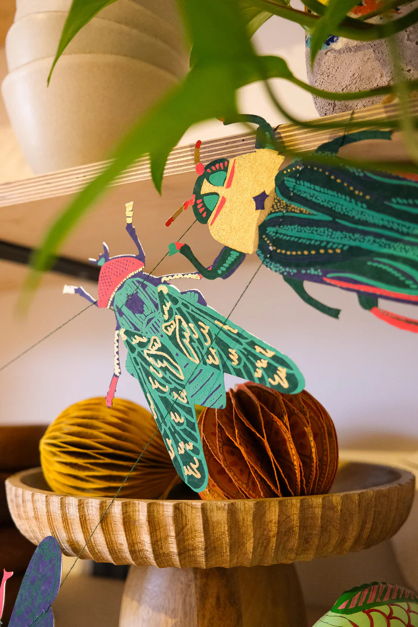 Vibrant paper insect garland featuring a large green moth and a gold and green beetle, hanging above a wooden bowl with