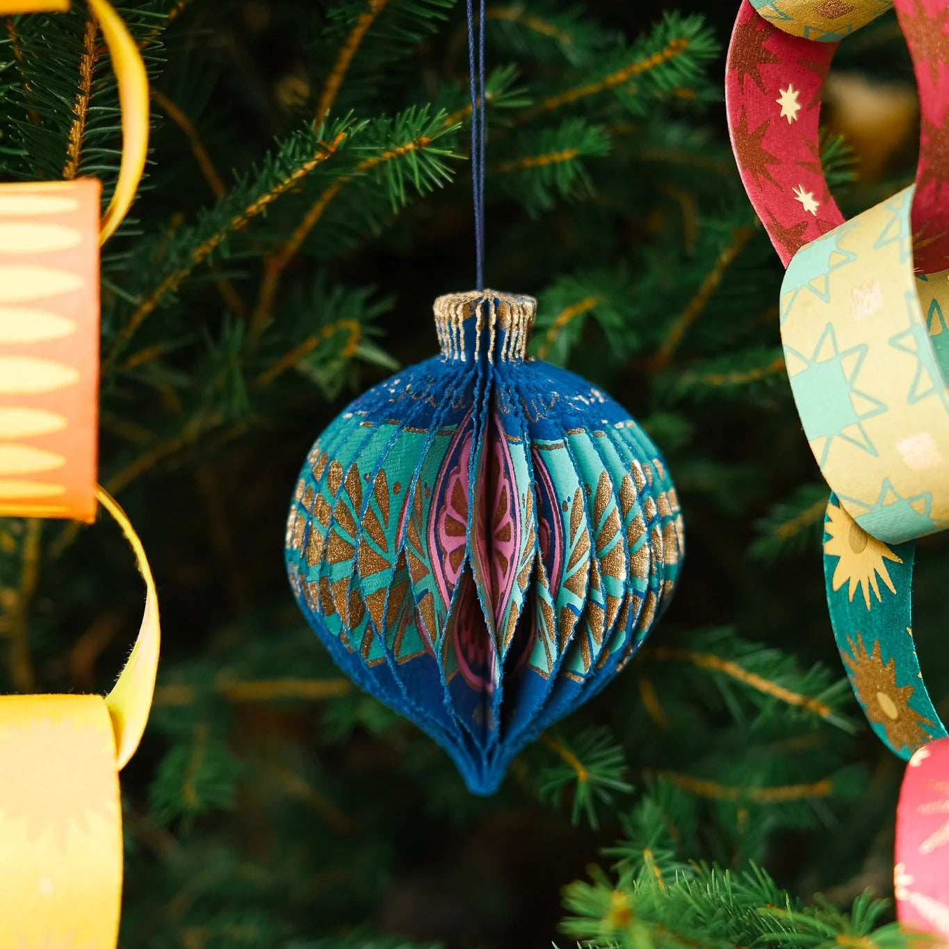 A decorative hanging ornament shaped like a folded paper teardrop in blue, teal, pink, and gold, suspended in front of green Christmas tree branches with colorful paper chains on both sides.