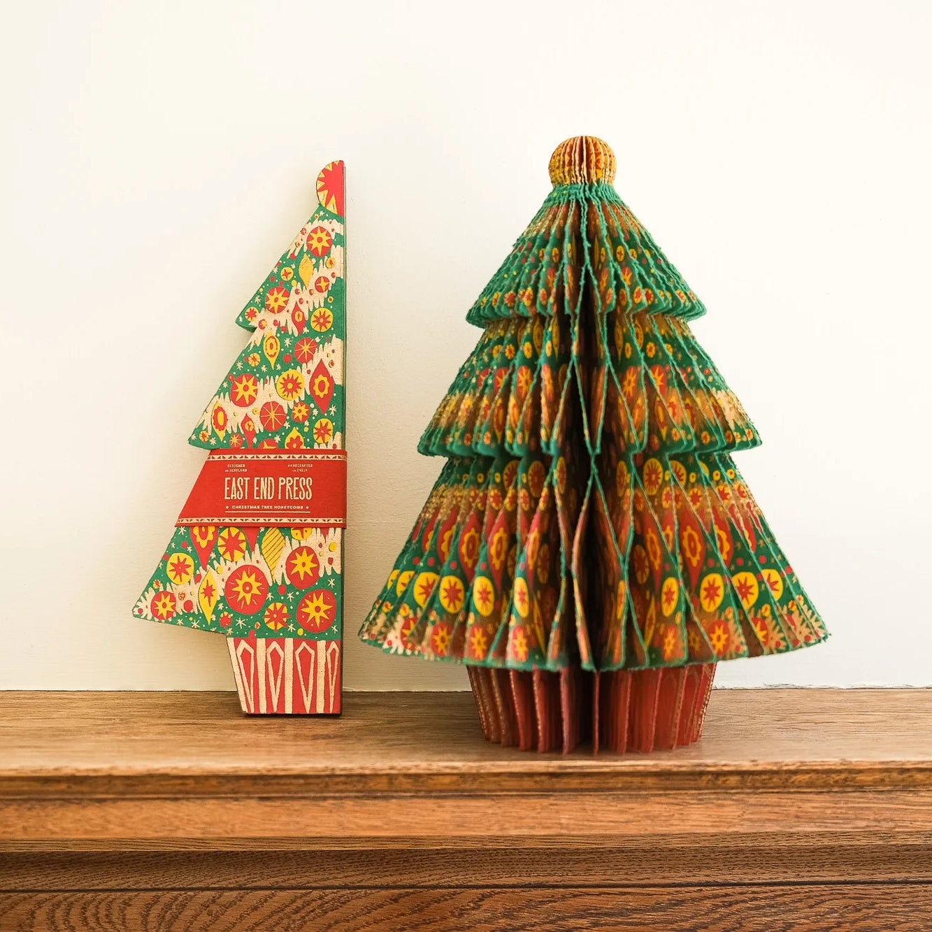Two decorative Christmas tree ornaments made from patterned paper, displayed on a wooden mantelpiece against a plain off-white wall.