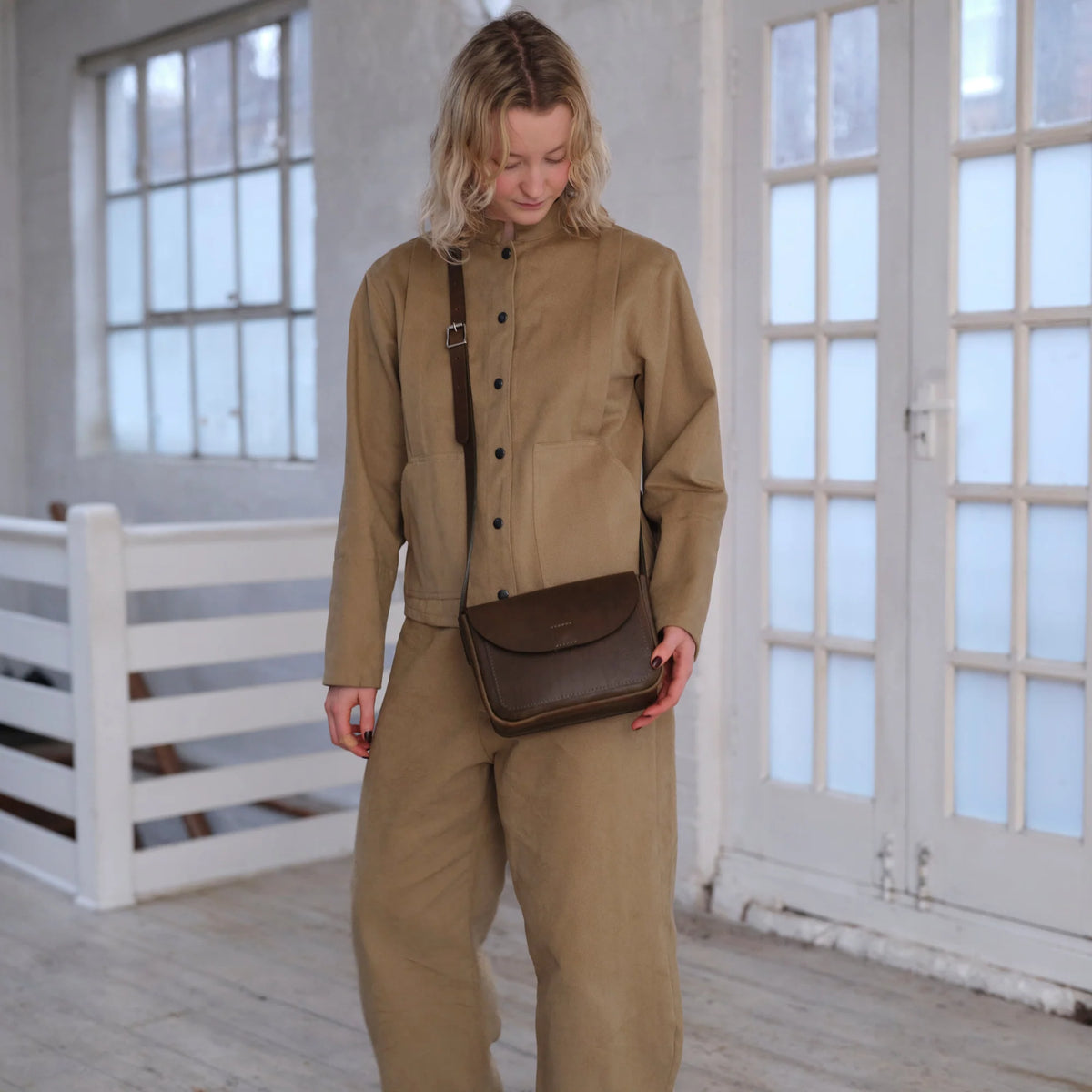 A woman wearing a matching beige corduroy jacket and trousers set, carrying a dark brown leather crossbody bag with a flap, standing in a bright room with white-framed windows.