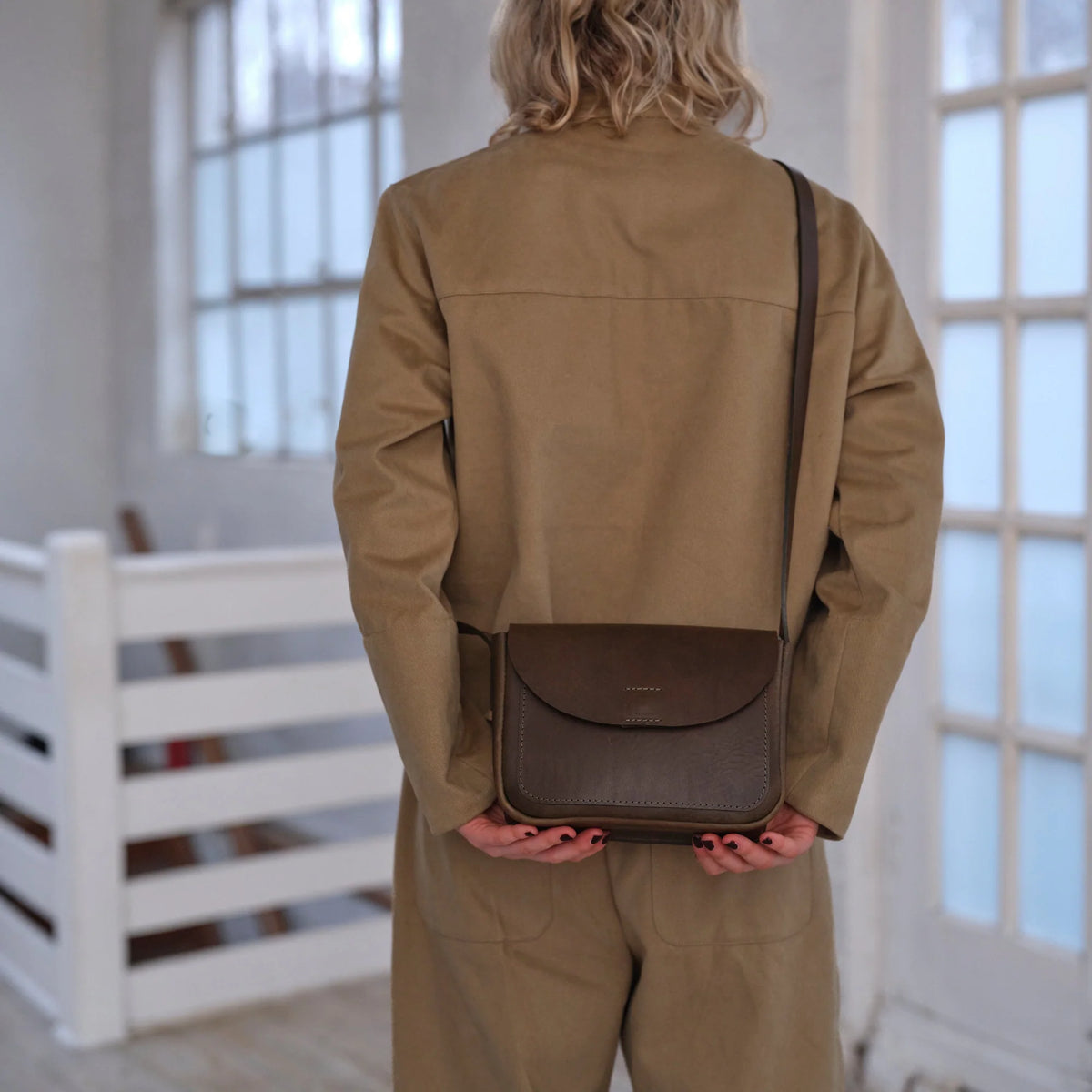 A person viewed from behind wearing a tan jumpsuit and holding a dark brown leather crossbody bag with a thin shoulder strap.