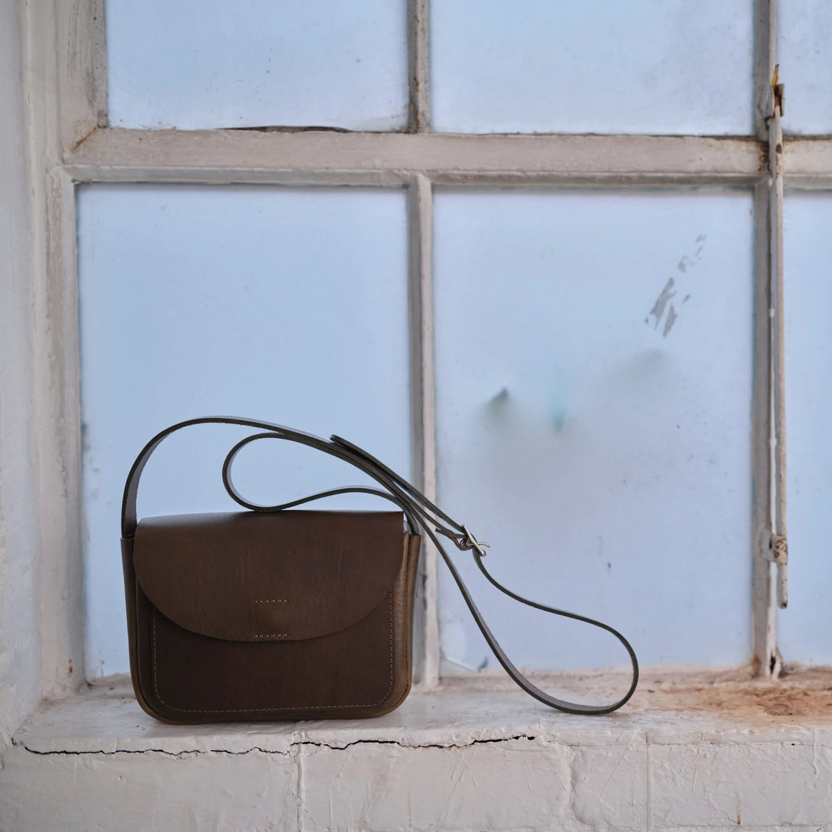 A dark brown leather crossbody bag with a long thin strap, resting on a white brick windowsill in front of a large white window.