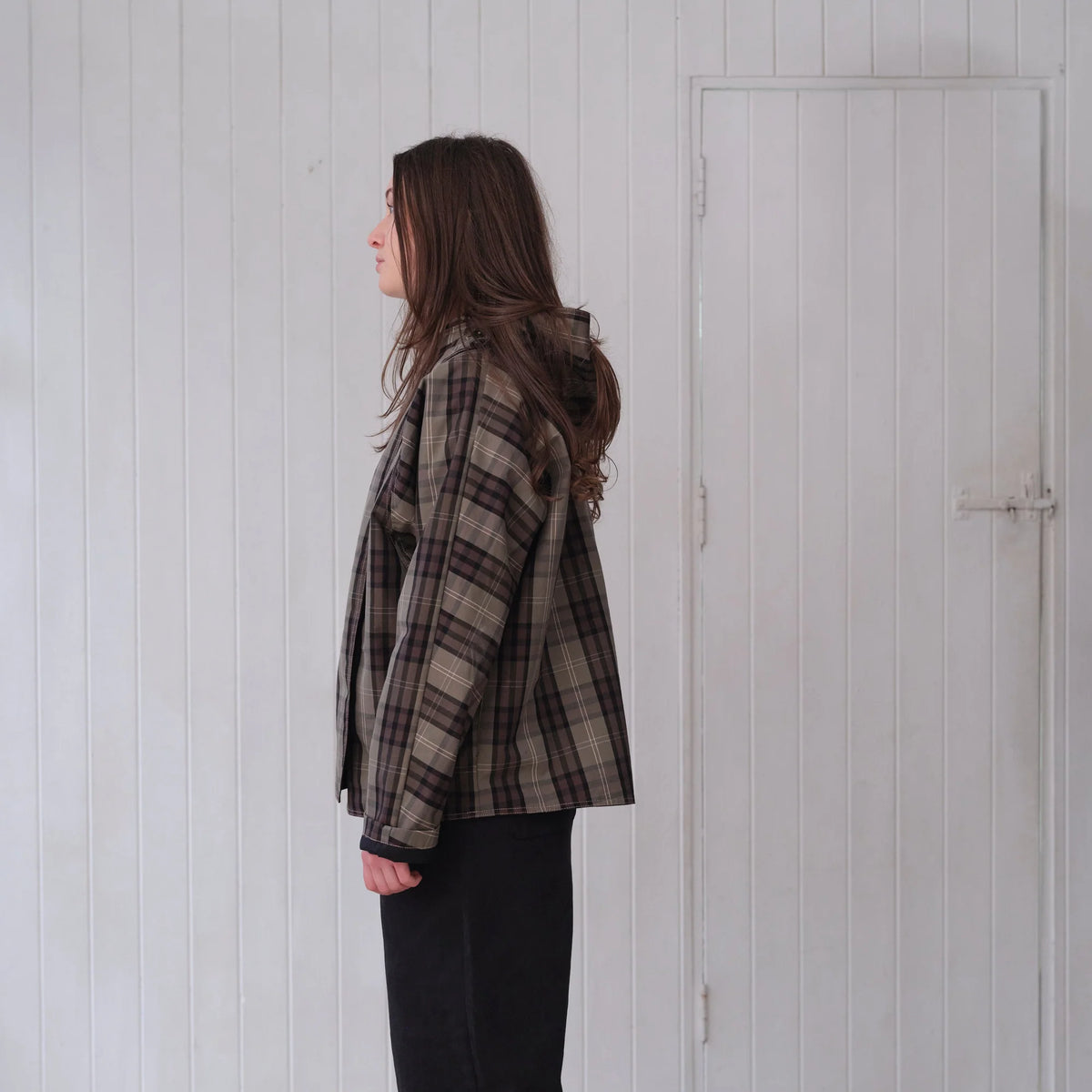 A woman stands in profile wearing a brown and beige plaid flannel shirt jacket with black pants, set against a white paneled wall and door.