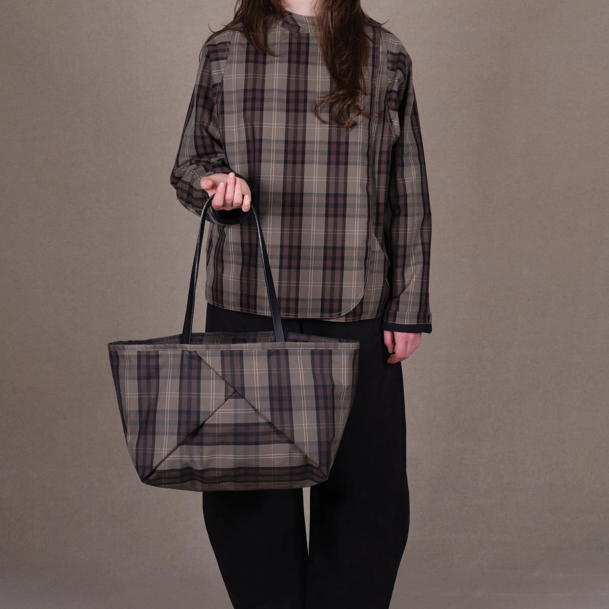 A person wearing a brown and black plaid long-sleeve shirt and black pants holds a matching plaid tote bag with black handles against a plain taupe background.
