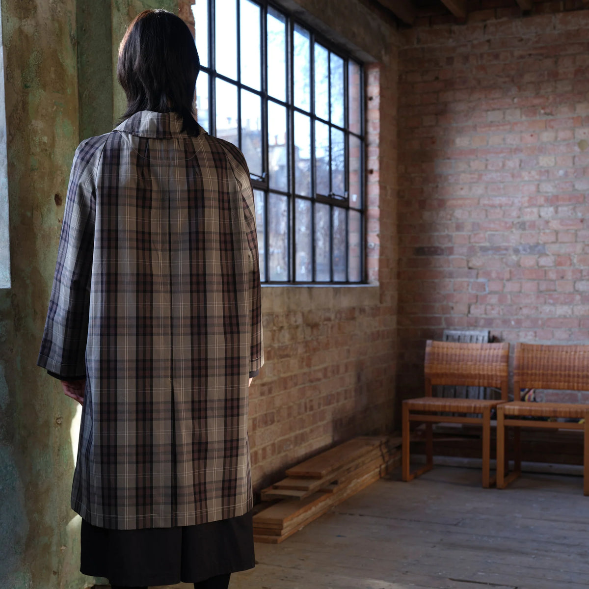 A person viewed from behind wearing a long beige and black plaid coat in a rustic brick room with a large window and two wooden chairs.